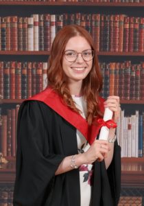 Siobhan is a white woman with red hair and glasses. This is a graduation picture in her black robes and red hood. 