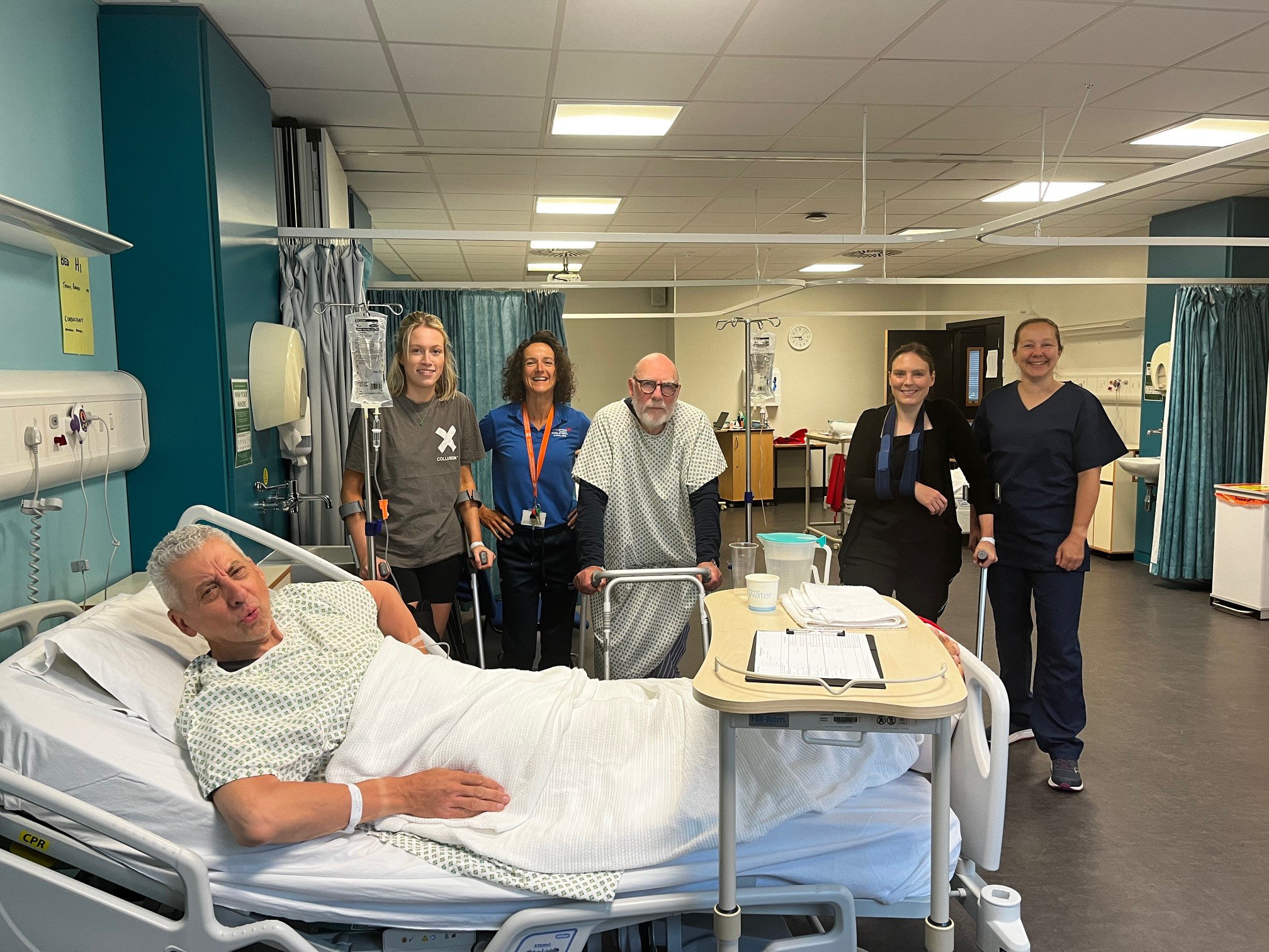 Director of Marketing & External Relations in a hospital bed in the simulation, and clinical skills centre, pretending to be in pain. He is surrounded by another simulated patient, students and staff. 