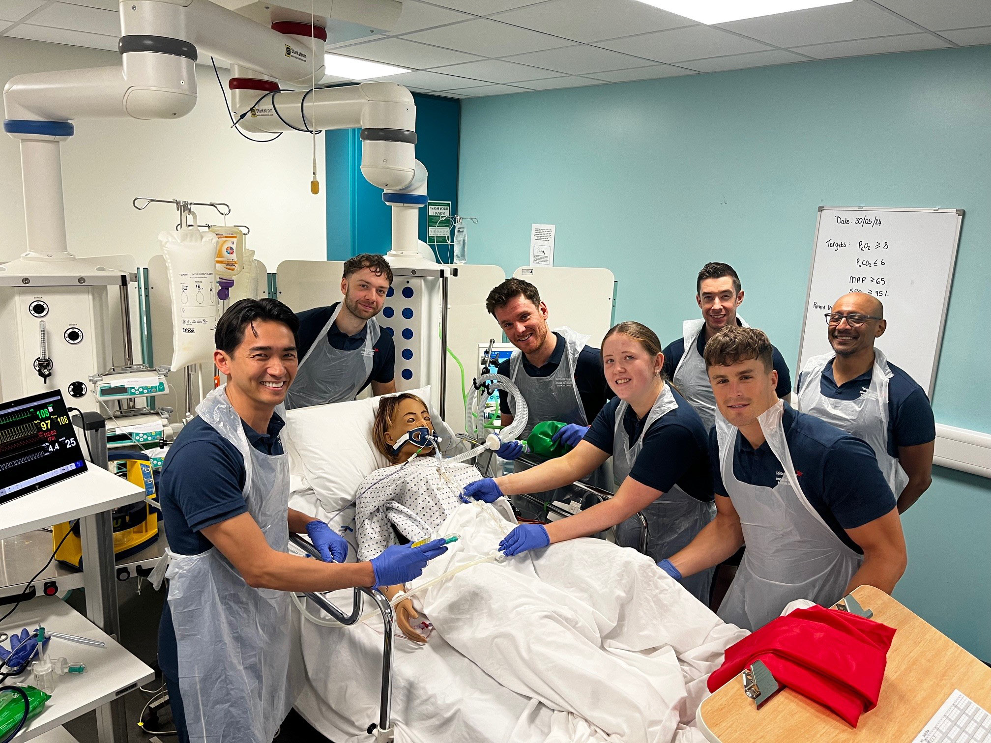 Students surrounding a mannequin which is connected to assisted breathing apparatus. The students are smiling and looking at the camera.