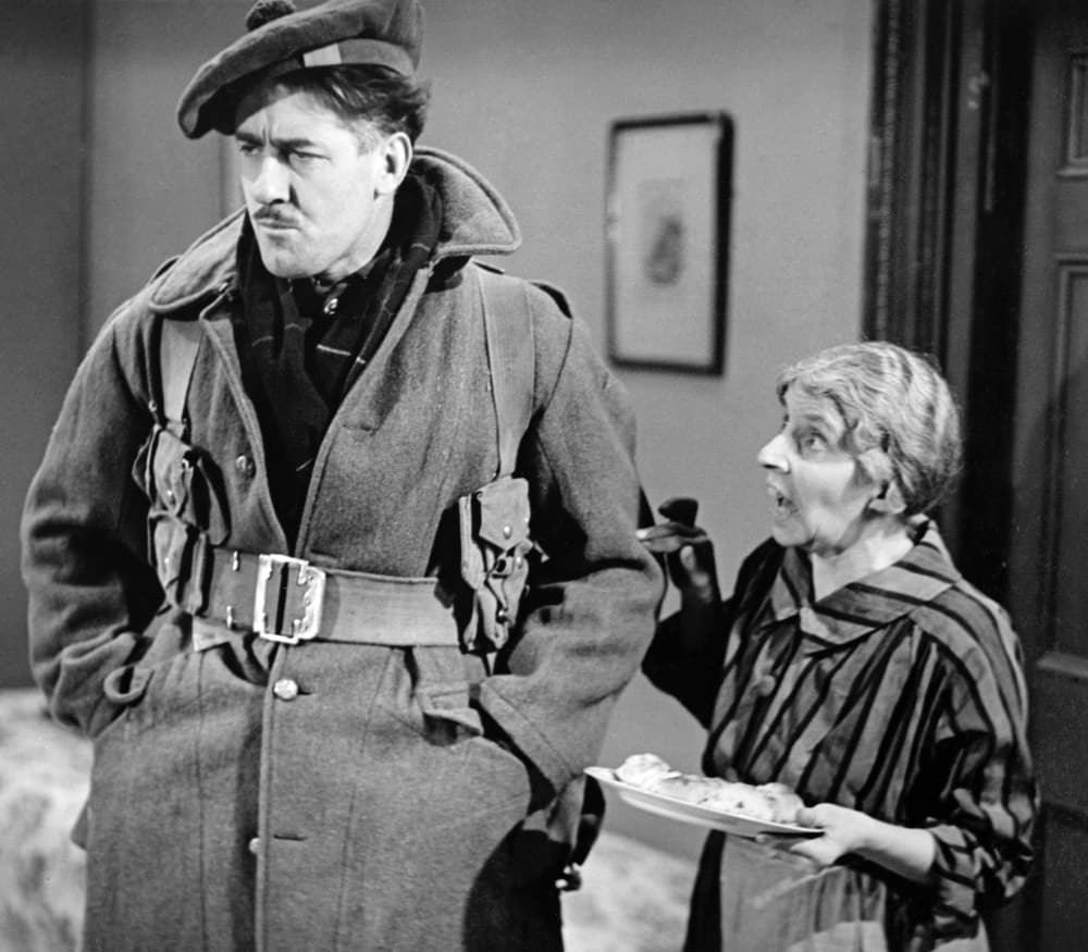 In a domestic scene, a tall military man in a greatcoat looks moodily offscreen to the left. An old woman talks animatedly to him, holding a plate.