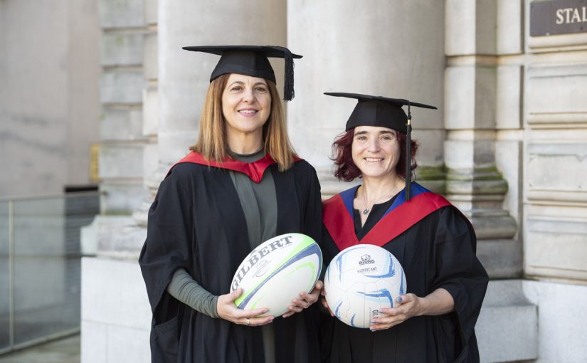 Christina Barrow and Clare Daniels standing outside Usher Hall in ther graduation gowns and hats, one of them holding a football and the other a rugby ball.
