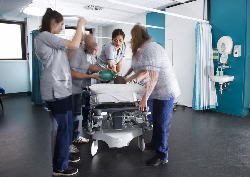 A slightly blurred image of a group of nurses in scrubs is seen giving life saving care with medical equipment to a patient on a hospital bed, in a clinical healthcare setting.