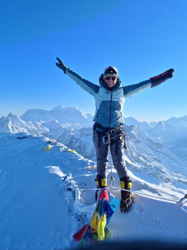 Devi is seen reaching the summit, with her arms extended in the air in their climbing gear. Snowy mountain terrain can be seen in the background with a clear blue sky.