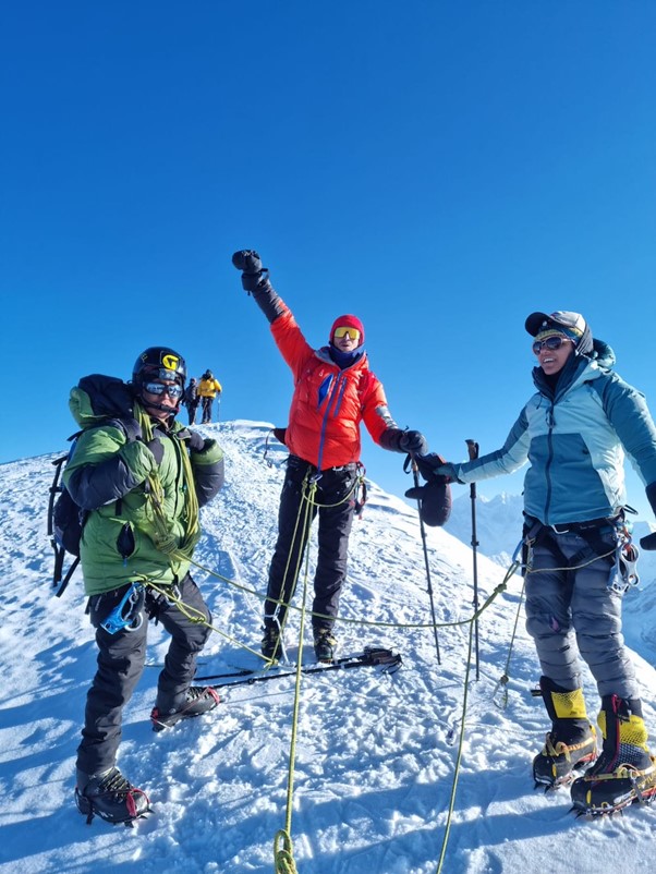 Members of the group are seen on their ascent. Snow is on the ground, with a mound seen behind them along with another two people in the shot, and a blue sky. One member has their hand in the air. 