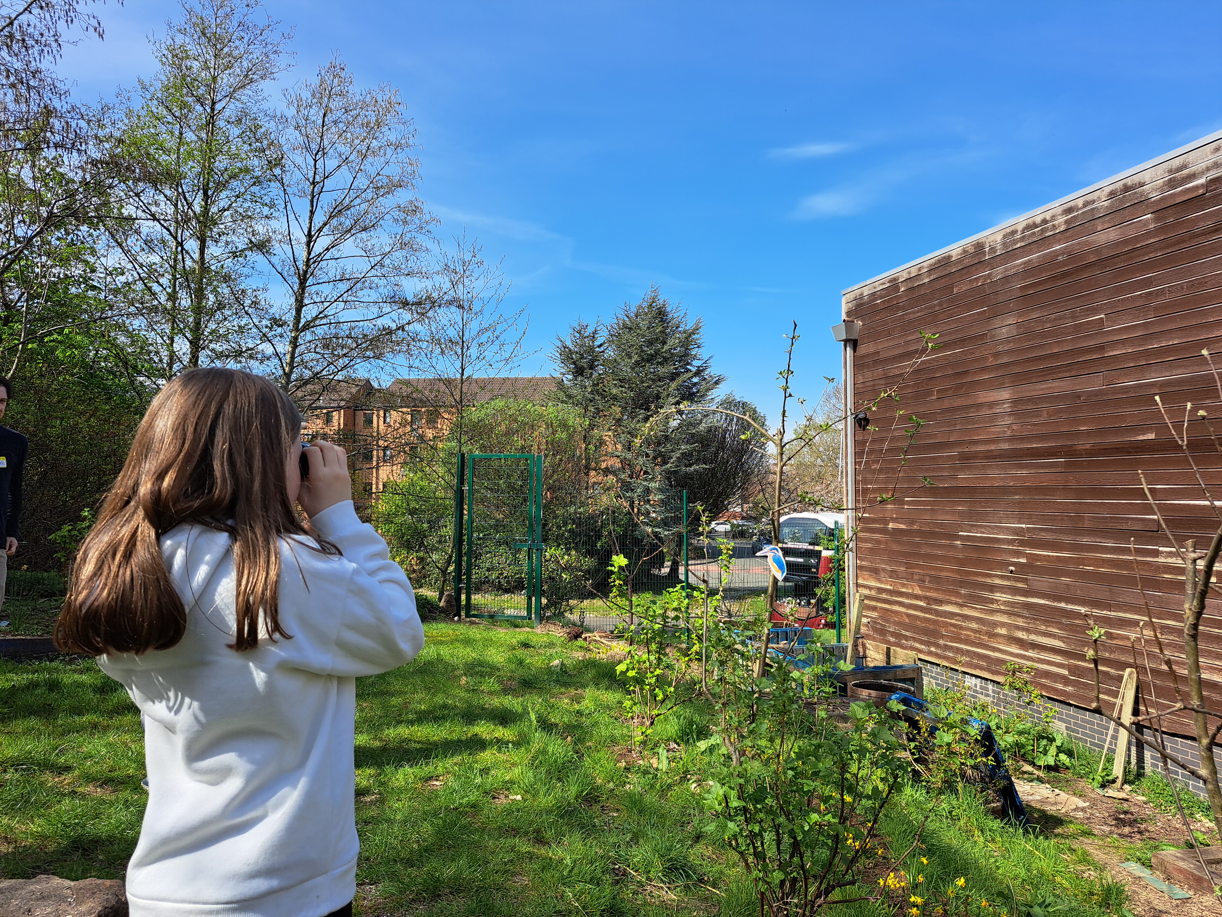 A girl stands with her back to us. She is looking through binoculars towards a bird in a garden.