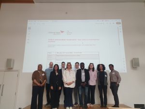 Ten people wearing smart casual clothes stand in front of a wall, with a projection of a powerpoint slide