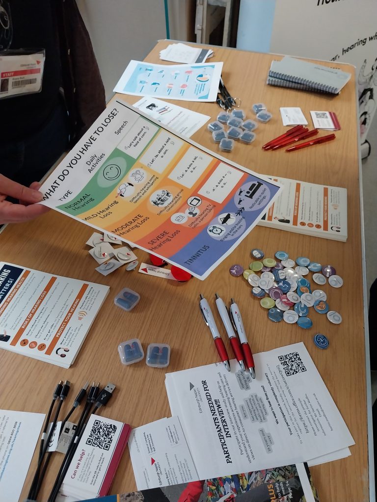 Table with leaflets about hearing loss, pens, ear plugs and badges. A person is handing out a leaflet over the table.