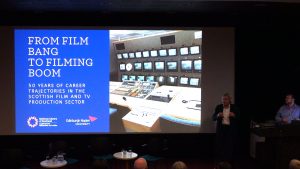 Picture shows a large screen showing a slide with title 'From 'Film Bang' to filming boom: Fifty years of career trajectories in the Scottish film and television production sector' and two men to the side presenting