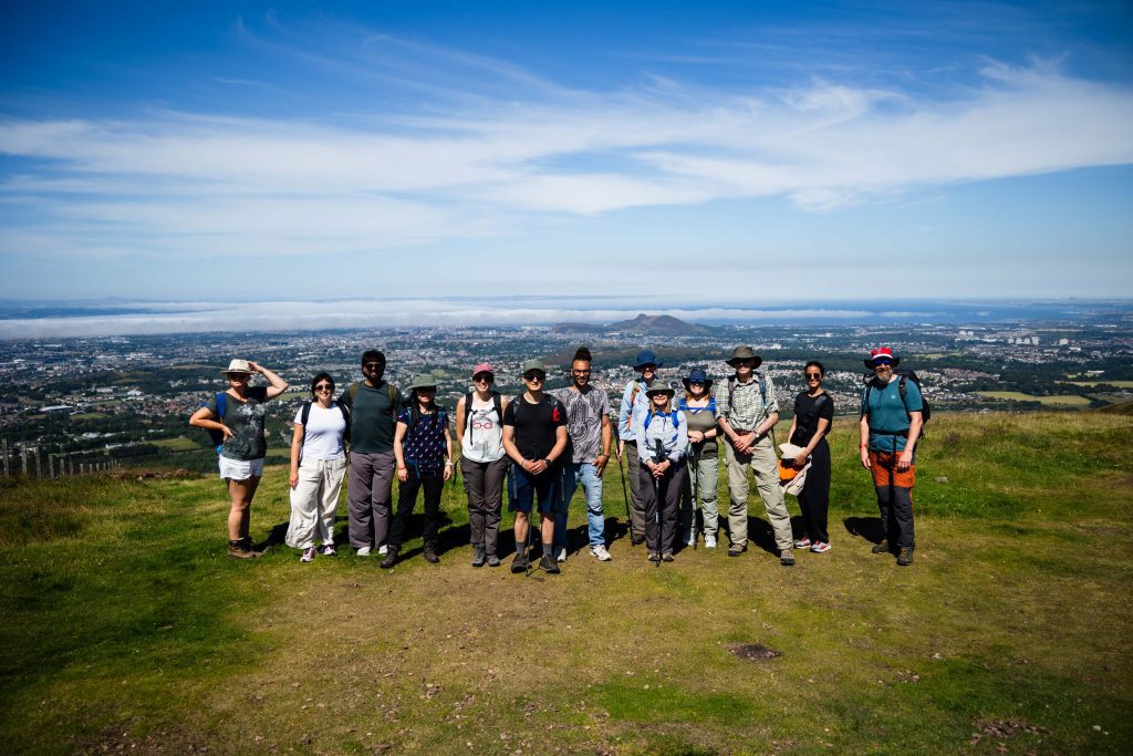 A group of 13 adults stand on a hill on a sunny day. In the background you can see the city of Edinburgh.