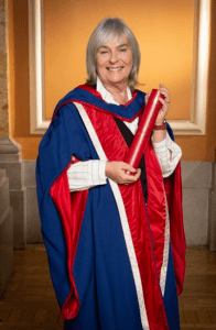 Susan Morrison, stands in front of a yellow background, dressed in Napier PhD graduation robes, holding a red scroll case.