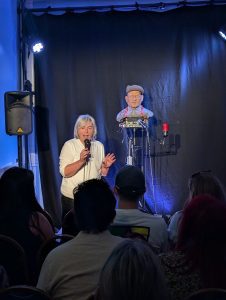 A woman stands in front of an audience holding a microphone. Behind her is a robot's head (which looks like an older white man) on a stand.
