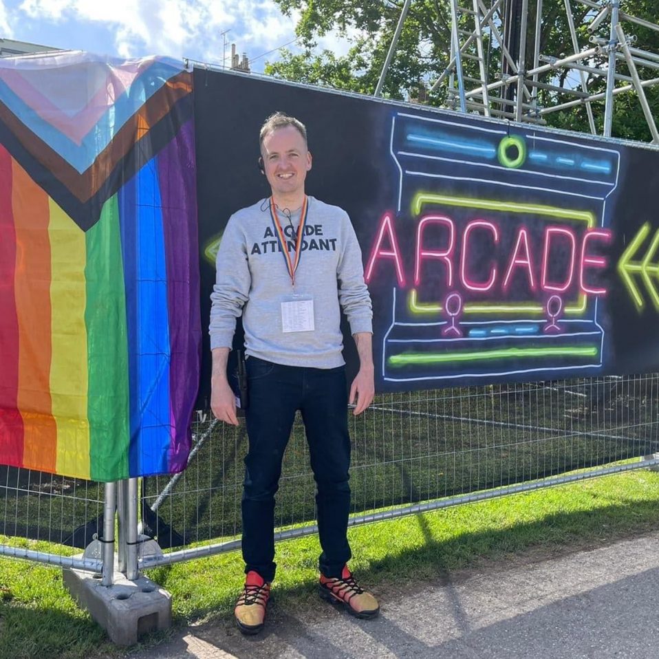 Dr Gary Kerr standing before a sign that says 'Arcade' and a two in one LGBTQAI+ and Transgender flag