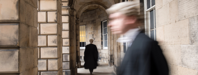 Blurred law practitioner in professional robe attire seen entering the image from the right, while a similar, focused image is seen walking through an archway further back.